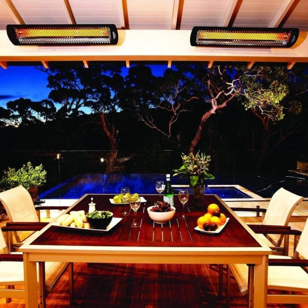 Outdoor dining area featuring a wooden table set with fresh fruits and salads, surrounded by a serene pool and lush trees, illuminated by stylish overhead heaters at dusk.