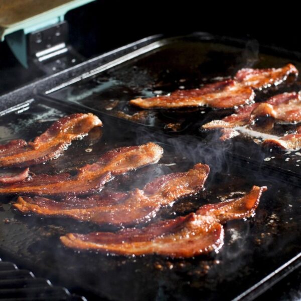 Crispy bacon strips sizzling on a griddle, releasing steam and cooking in their own grease.