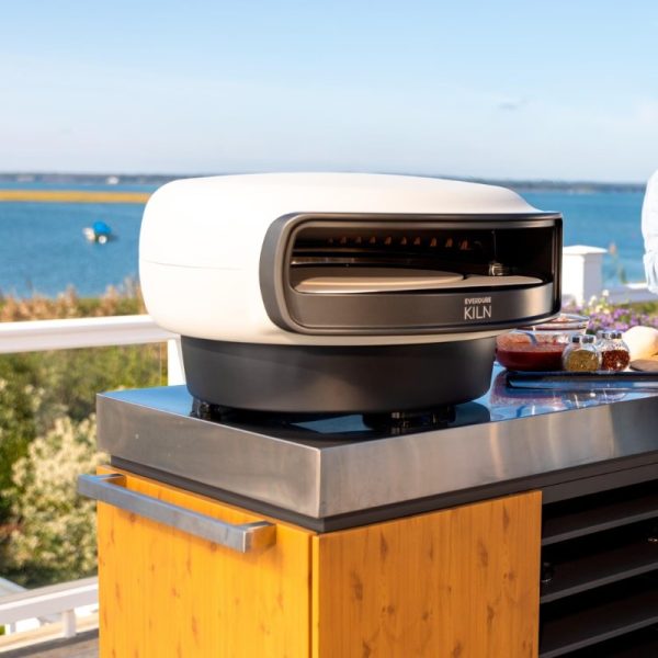 Everdure Kiln outdoor pizza oven on a countertop with scenic waterfront background, garnished with ingredients for cooking.