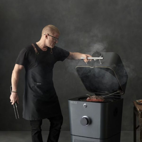 Man in an apron using tongs to check a piece of meat on a modern grill, surrounded by smoke, showcasing outdoor cooking techniques.