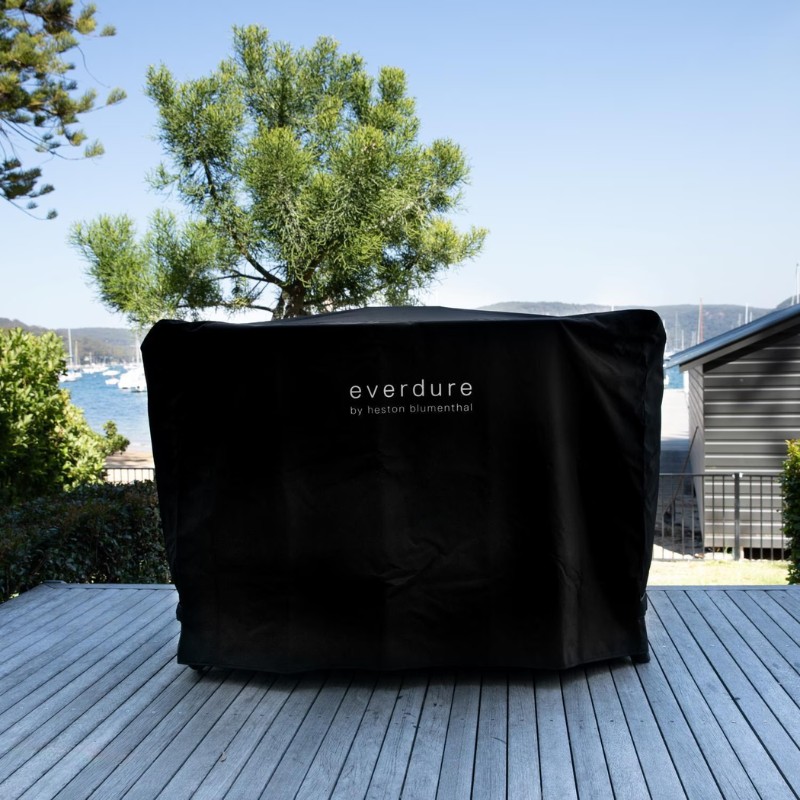Black protective cover for an Everdure barbecue by Heston Blumenthal, set on a wooden deck with a scenic view of the water and greenery.
