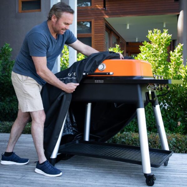 Man removing a cover from an orange Everdure barbecue grill on a wooden deck surrounded by greenery.