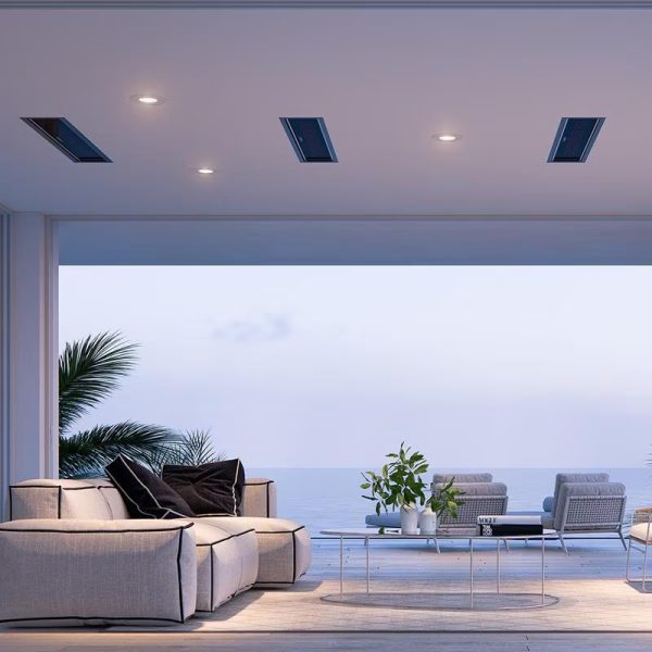 Modern coastal living room with a view of the ocean, featuring a light-colored sectional sofa, a glass coffee table, and potted plants, illuminated by recessed ceiling lights.