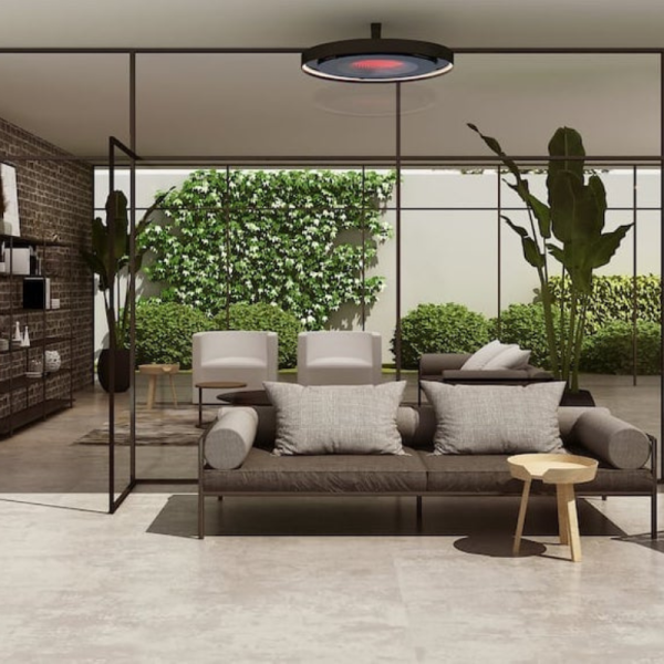 Modern living room with a dark sofa and light-colored armchairs, featuring large glass walls that open to a garden with greenery. A stylish pendant light hangs above, and a small wooden side table complements the decor.