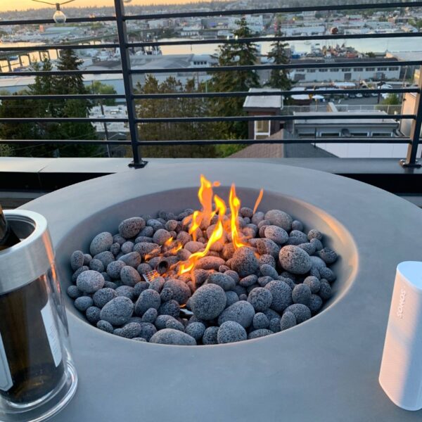 Modern outdoor fire pit with glowing flames surrounded by smooth gray stones, overlooking a scenic waterfront view at sunset, featuring a wine bottle and portable speaker nearby.