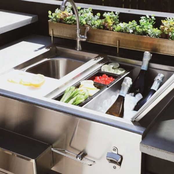 Outdoor kitchen sink with integrated ice compartment, featuring fresh ingredients like lemons, tomatoes, and celery, along with chilled bottles of champagne.