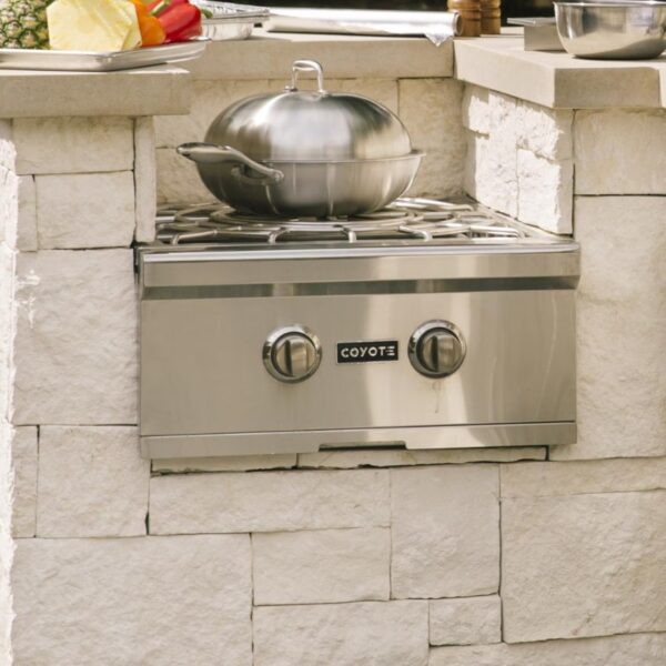 Stainless steel outdoor cooking station with a Coyote gas burner and a covered pot, set against a stone wall.