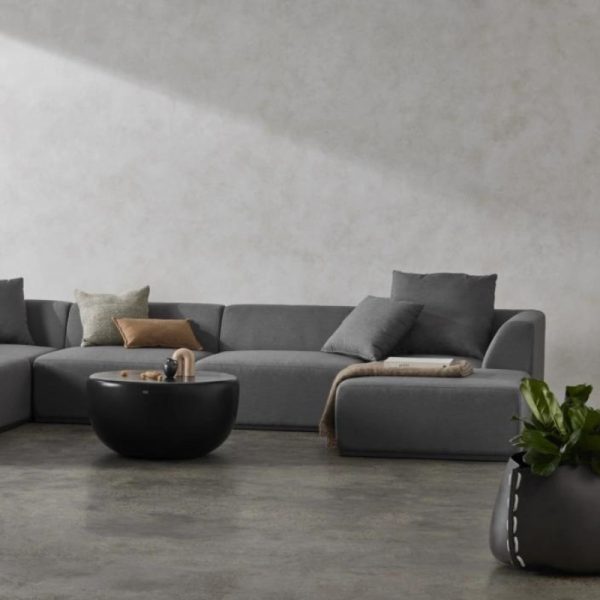 Modern gray sectional sofa with decorative cushions, accompanied by a round black coffee table and a potted plant, set against a textured light wall.
