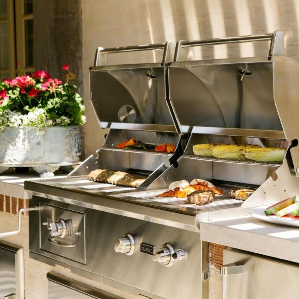 Stainless steel outdoor grill with various meats and vegetables cooking, surrounded by potted flowers. Ideal for summer barbecues and outdoor cooking.