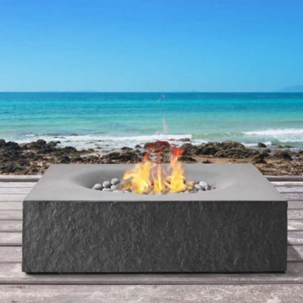 Modern outdoor fire pit on a wooden deck by the beach, with flames and decorative stones, against a backdrop of blue ocean and clear sky.