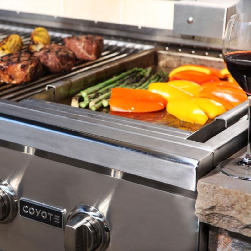 Close-up of a Coyote grill with grilled meats, asparagus, and colorful bell peppers cooking on a stainless steel surface, accompanied by a glass of red wine.