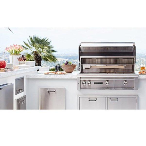 Stainless steel outdoor kitchen grill with a marble countertop, surrounded by plants and dishes, overlooking a scenic view.
