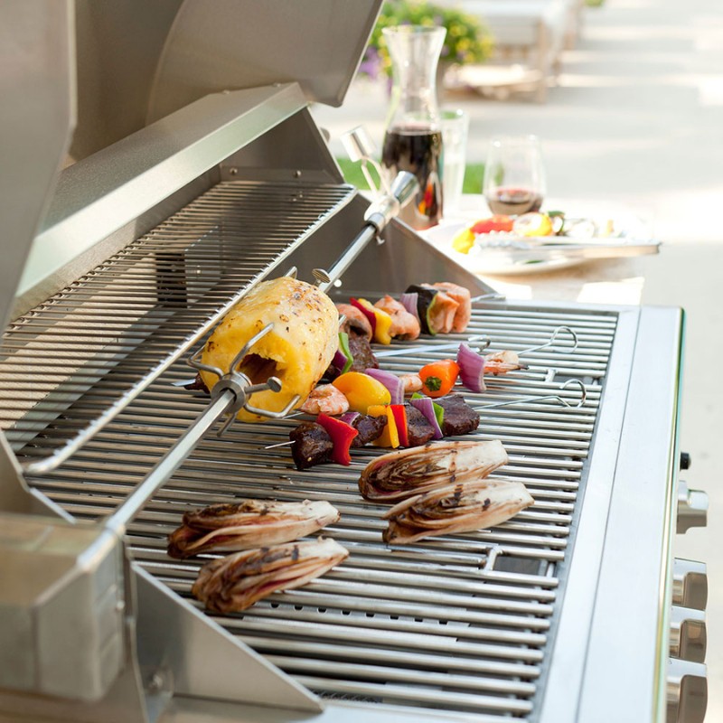 Grilled meats and vegetables on a stainless steel barbecue grill, featuring a rotisserie with a whole chicken and colorful skewers, set in a backyard with drinks and appetizers in the background.