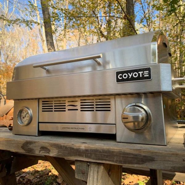 Stainless steel Coyote grill on a wooden table in a forest setting, showcasing its sleek design and control knobs.