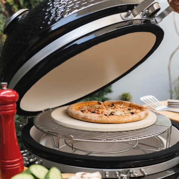 Pizza cooking on a ceramic grill with the lid open, showcasing a golden crust and toppings, surrounded by fresh ingredients like cucumbers and a red pepper grinder.