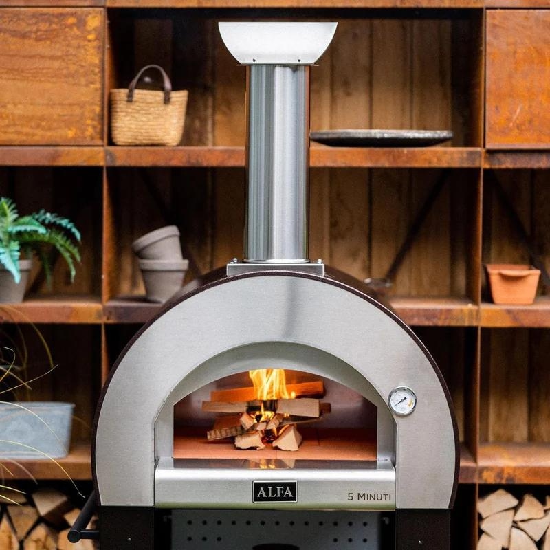 Alfa wood-fired pizza oven with flames visible inside, set against a rustic wooden shelf with plants and pottery. Ideal for outdoor cooking and entertaining.