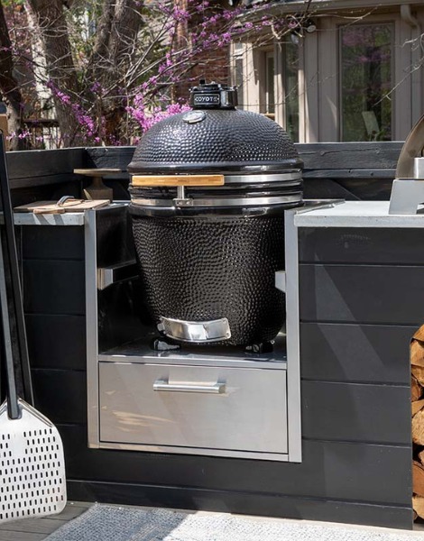 Black ceramic grill in an outdoor kitchen setup, featuring stainless steel storage below and surrounded by wooden accents.