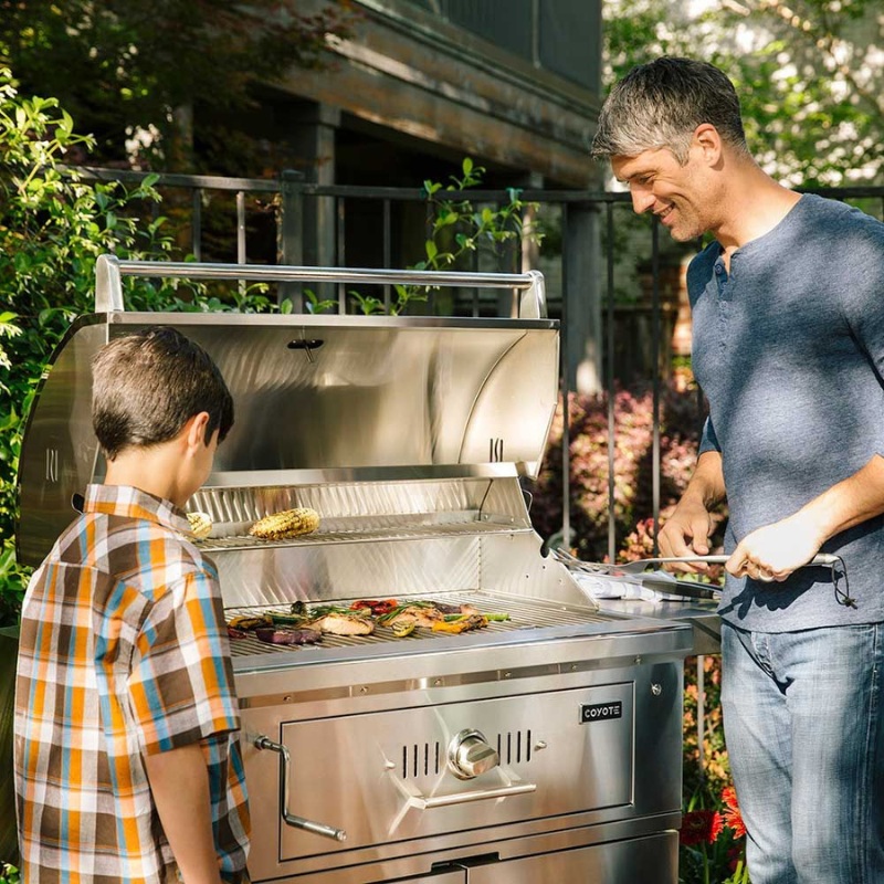 Father and son grilling vegetables and corn on a stainless steel Coyote grill in a backyard setting.