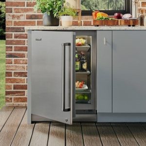 Modern outdoor kitchen with a stainless steel refrigerator, partially open to reveal drinks and fresh produce, against a brick wall.