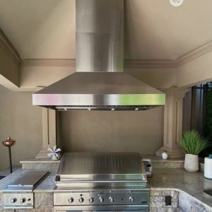 Stainless steel outdoor kitchen with a modern hood and grill setup, featuring granite countertops and decorative plants.