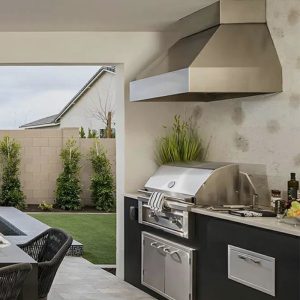 Modern outdoor kitchen featuring a stainless steel grill and hood, surrounded by greenery and a dining area with wicker chairs.