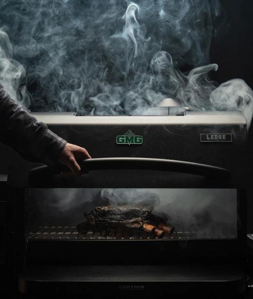 Hand opening the lid of a GMG Ledge grill, with smoke billowing out and a piece of meat visible inside, showcasing outdoor cooking and grilling.