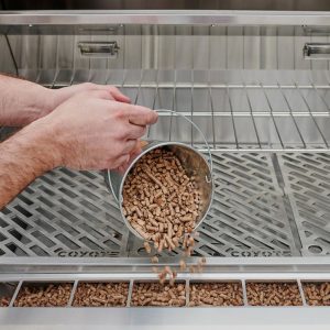 Hands pouring wood pellets from a metal bucket into a stainless steel grill, showcasing the grill's interior design and functionality.