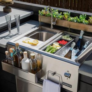 Outdoor kitchen sink setup featuring stainless steel sink, fresh ingredients like lemons and limes, drinks on ice, and elegant glassware, perfect for entertaining.