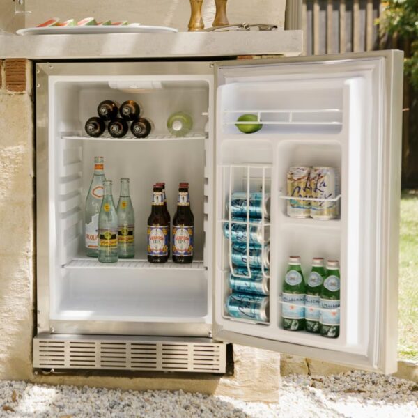 Outdoor refrigerator stocked with various beverages, including bottled water, craft beers, and soda cans, set against a sunny backyard backdrop.