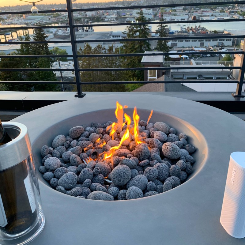 Modern rooftop fire pit surrounded by smooth stones, with a bottle of wine and a wireless speaker nearby, overlooking a scenic waterfront view at sunset.