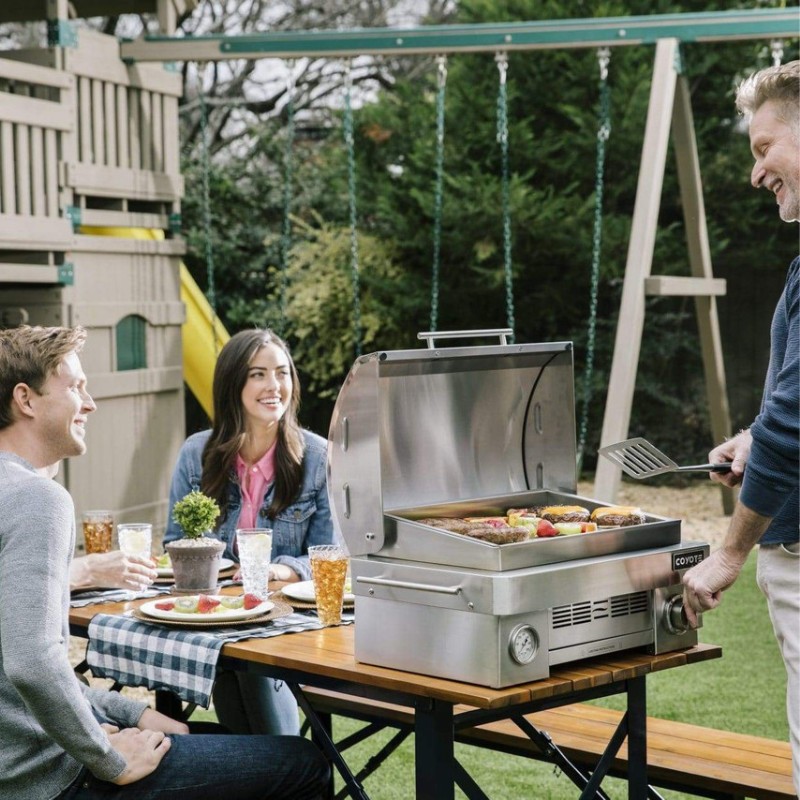 Group of friends enjoying a backyard barbecue, with a stainless steel grill cooking burgers and vegetables, seated at a picnic table with drinks and plates of food.