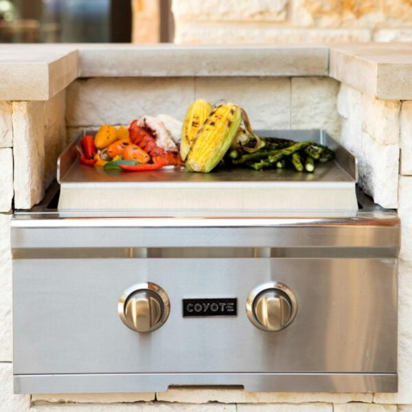 Stainless steel Coyote outdoor grill with grilled vegetables, corn, and lobster on a stone patio.