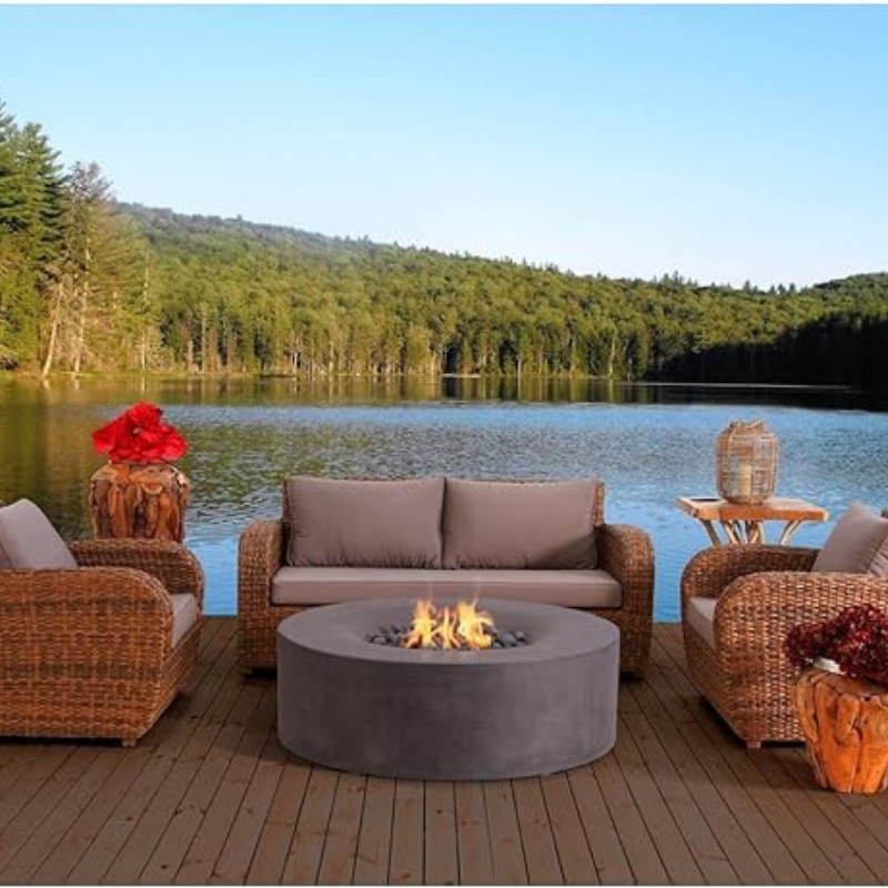 Cozy outdoor seating area by a serene lake, featuring a circular fire pit, wicker furniture, and lush green trees in the background.