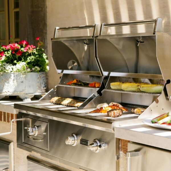 Stainless steel outdoor grill with various grilled meats and vegetables, surrounded by vibrant flowers in a decorative planter.