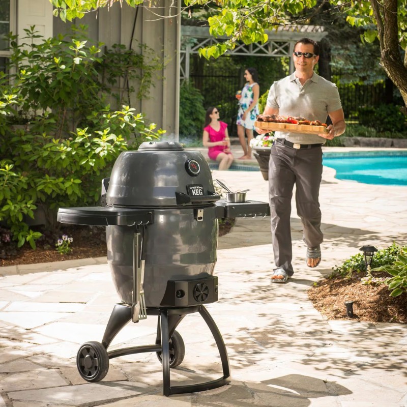 Man carrying a tray of food near a gray outdoor grill by a pool, with two women enjoying drinks in the background, surrounded by greenery.