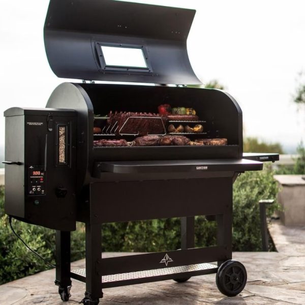 Black pellet grill with an open lid showcasing various meats and vegetables on the cooking racks, set in an outdoor environment with greenery.