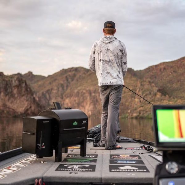 Person fishing from a boat on a calm lake, surrounded by mountains, with fishing gear and a grill visible on the deck.