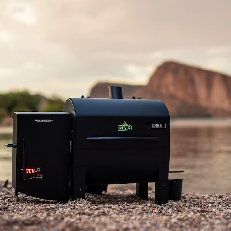GMG Trek grill positioned by a scenic lakeside, showcasing its sleek black design and digital temperature display. Ideal for outdoor cooking and smoking.