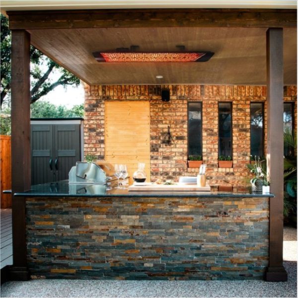 Outdoor kitchen with a stone bar, modern appliances, and a rustic brick wall backdrop, featuring large windows and decorative plants.
