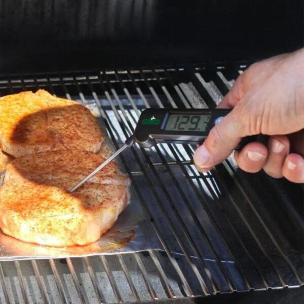 Hand using a digital meat thermometer to check the internal temperature of seasoned pork chops on a grill, displaying a reading of 129.3°F.