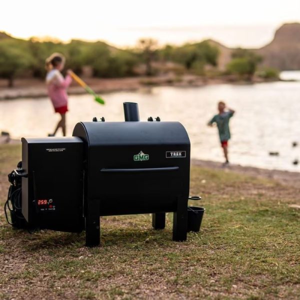 GMG Trek pellet grill positioned on the grass near a lake, with children playing in the background. Ideal for outdoor cooking and family gatherings.