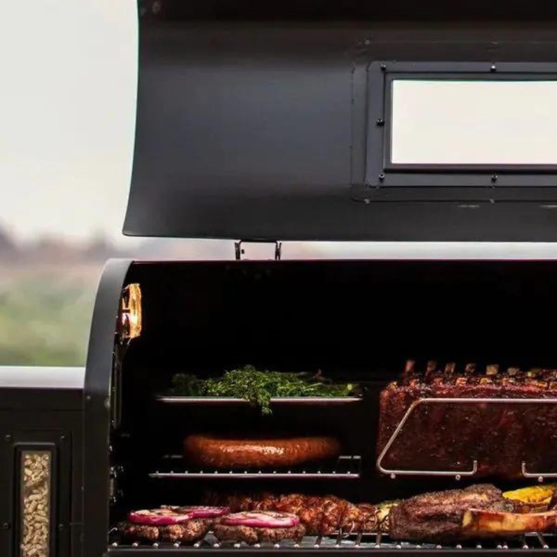 Close-up of a barbecue grill with various meats, including steaks and sausages, being smoked alongside fresh herbs, showcasing a delicious grilling setup.