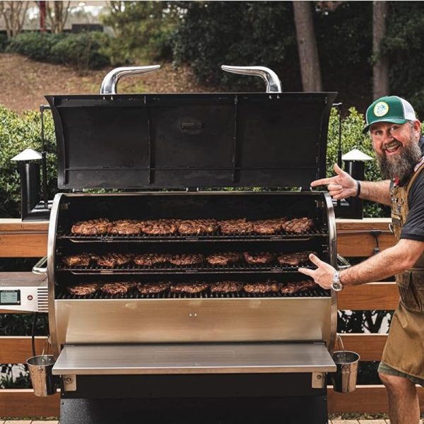Man proudly showcasing a fully loaded smoker grill with multiple racks of perfectly cooked meat, set in a backyard setting.