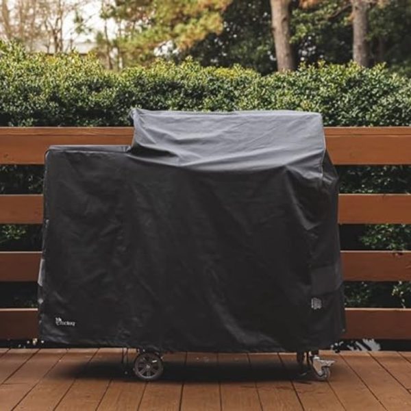 Black grill cover on a wooden deck, surrounded by greenery, protecting the grill from the elements.