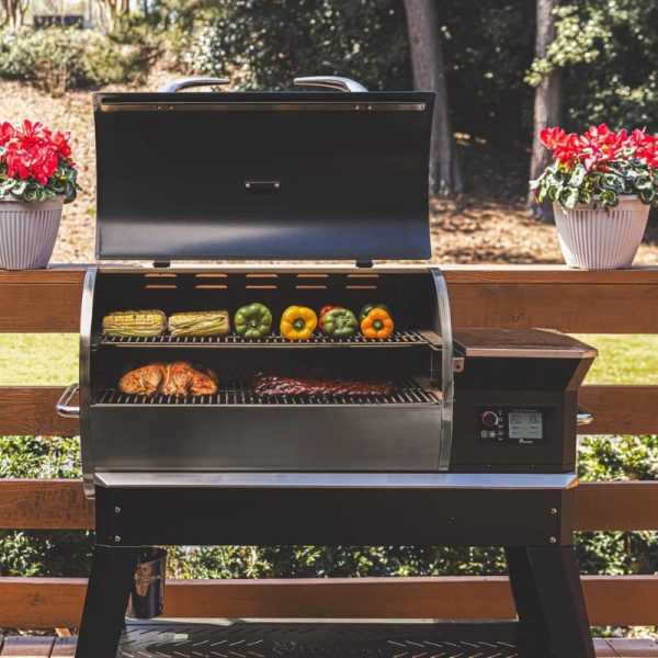 Outdoor grill with meats and vegetables cooking on the rack, featuring a black exterior, open lid, and surrounded by potted flowers on a wooden deck.