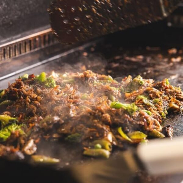 Sizzling stir-fried vegetables and meat on a hot grill, with steam rising and a spatula in action.