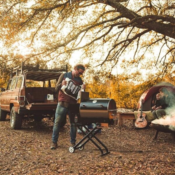 Man preparing a grill at a campsite with a vintage truck in the background, surrounded by autumn foliage and a cozy seating area.