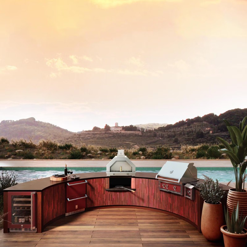 Outdoor kitchen featuring a curved wooden countertop, built-in grill, and wine cooler, with a scenic view of hills and a pool at sunset.