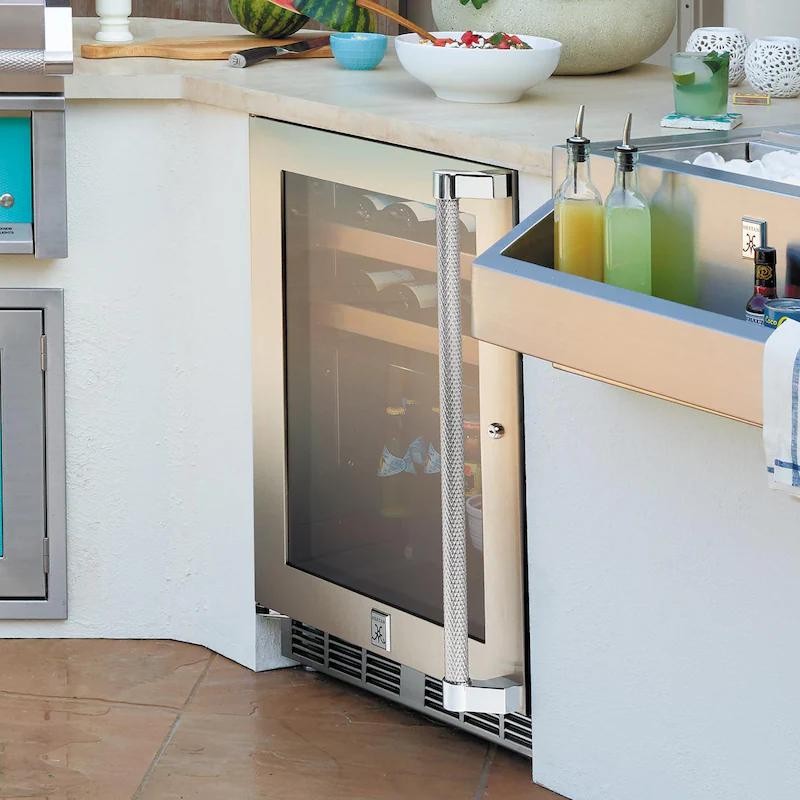 Outdoor kitchen featuring a stainless steel wine cooler with a glass door, adjacent to a beverage station with colorful drinks and ice.
