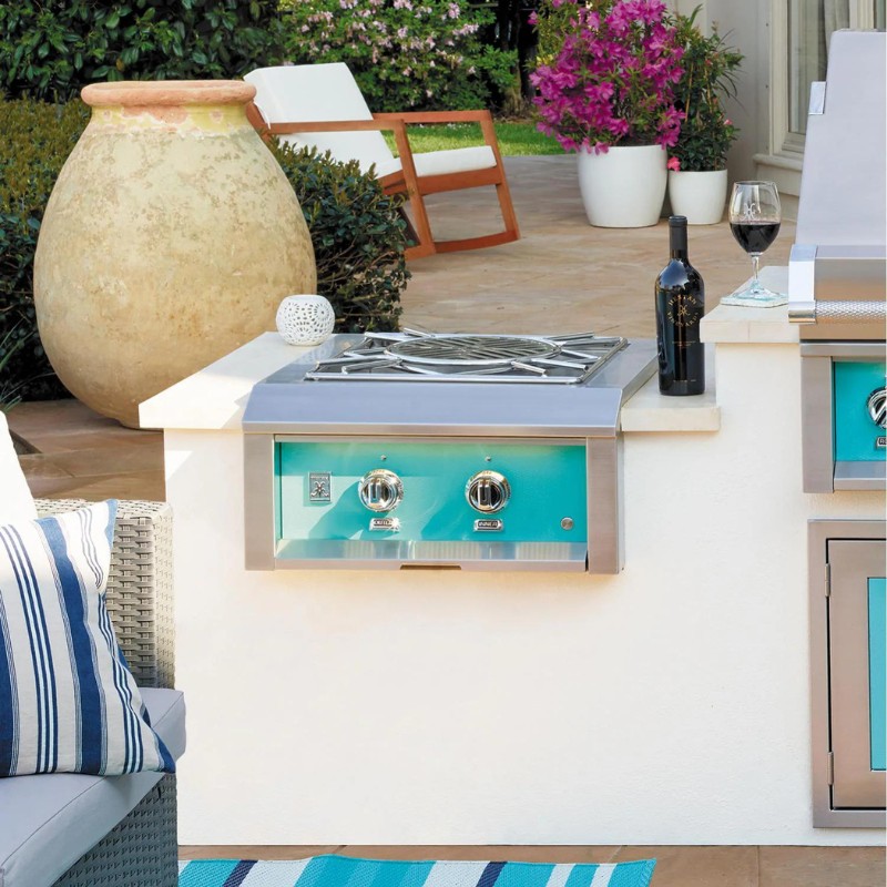 Outdoor kitchen featuring a stainless steel gas grill with turquoise accents, surrounded by a cozy patio setting with a wine bottle and glass, decorative pottery, and lush greenery.